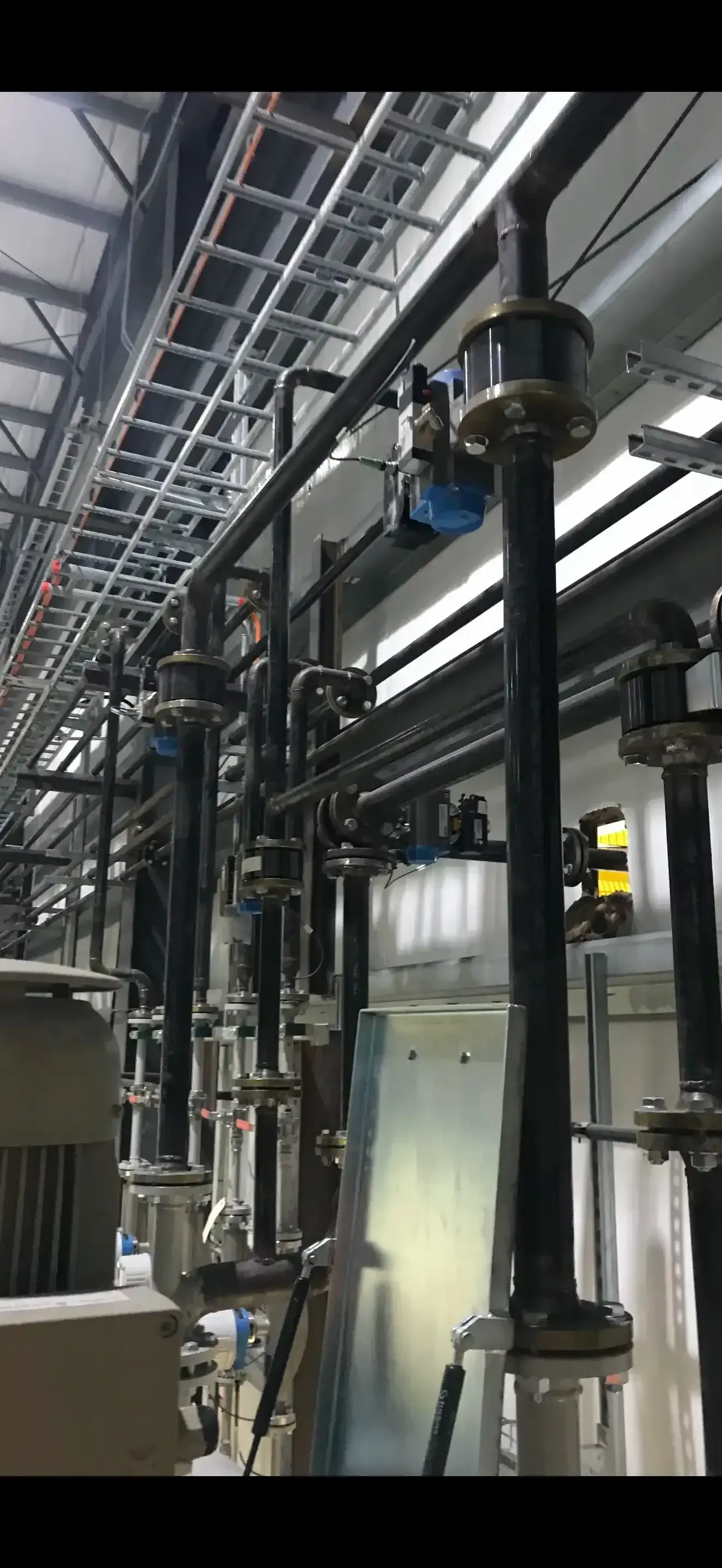 Industrial equipment with black pipes, silver fixtures, and overhead cable trays in a large factory.