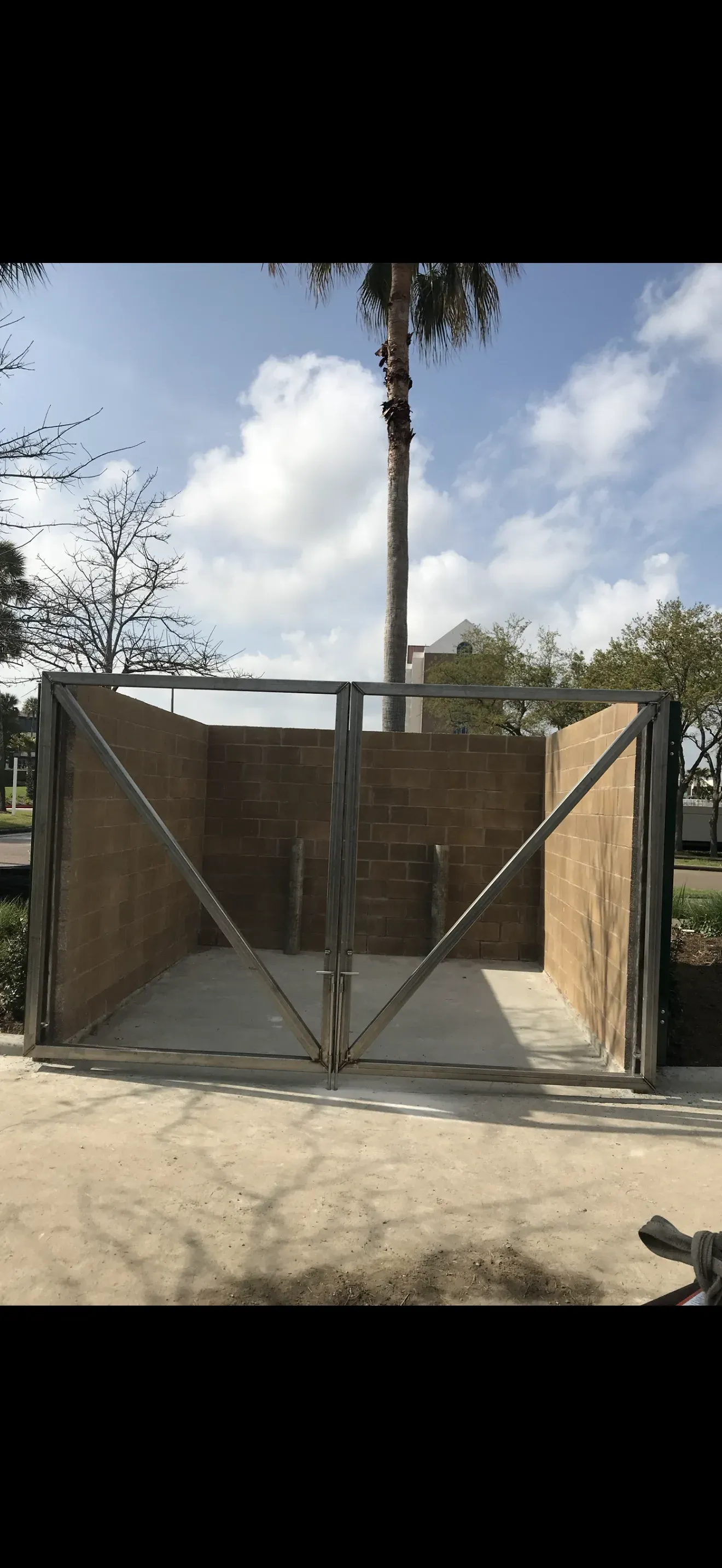 Steel-framed dumpster enclosure gate on a concrete pad, palm trees and blue sky behind.