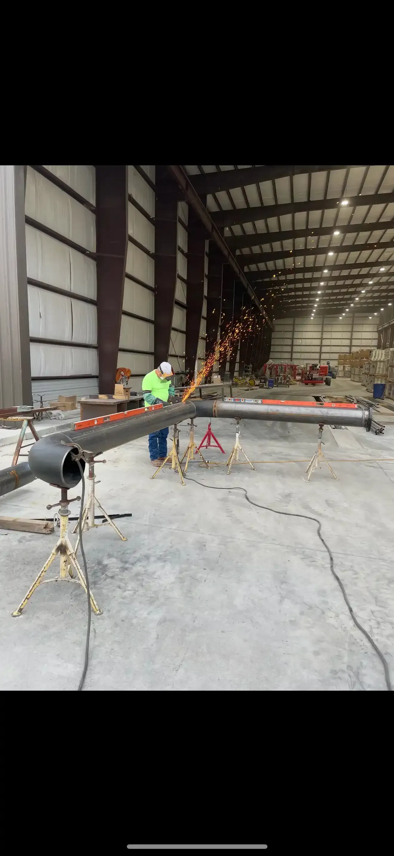Welder cutting a long steel pipe set on pipe stands, sparks flying, inside a large fabrication shop.